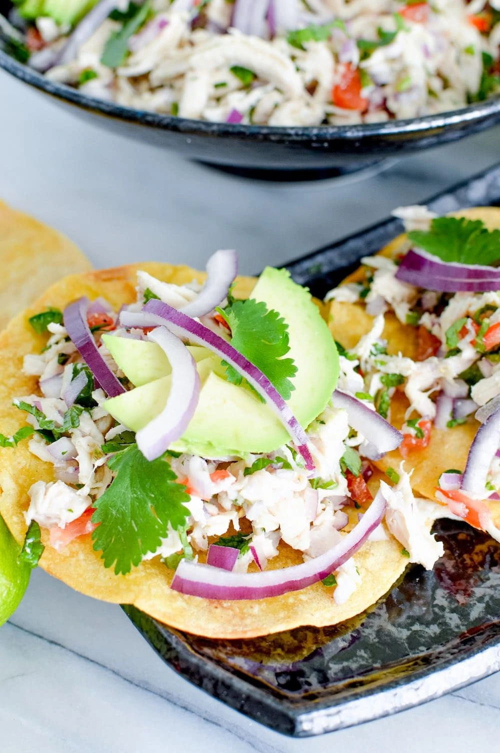 Two tostadas topped with shredded chicken, sliced avocado, red onion, cilantro, and a tomato mixture are served on a black dish with more filling in the background—perfect for summer recipes or cold chicken recipes.
