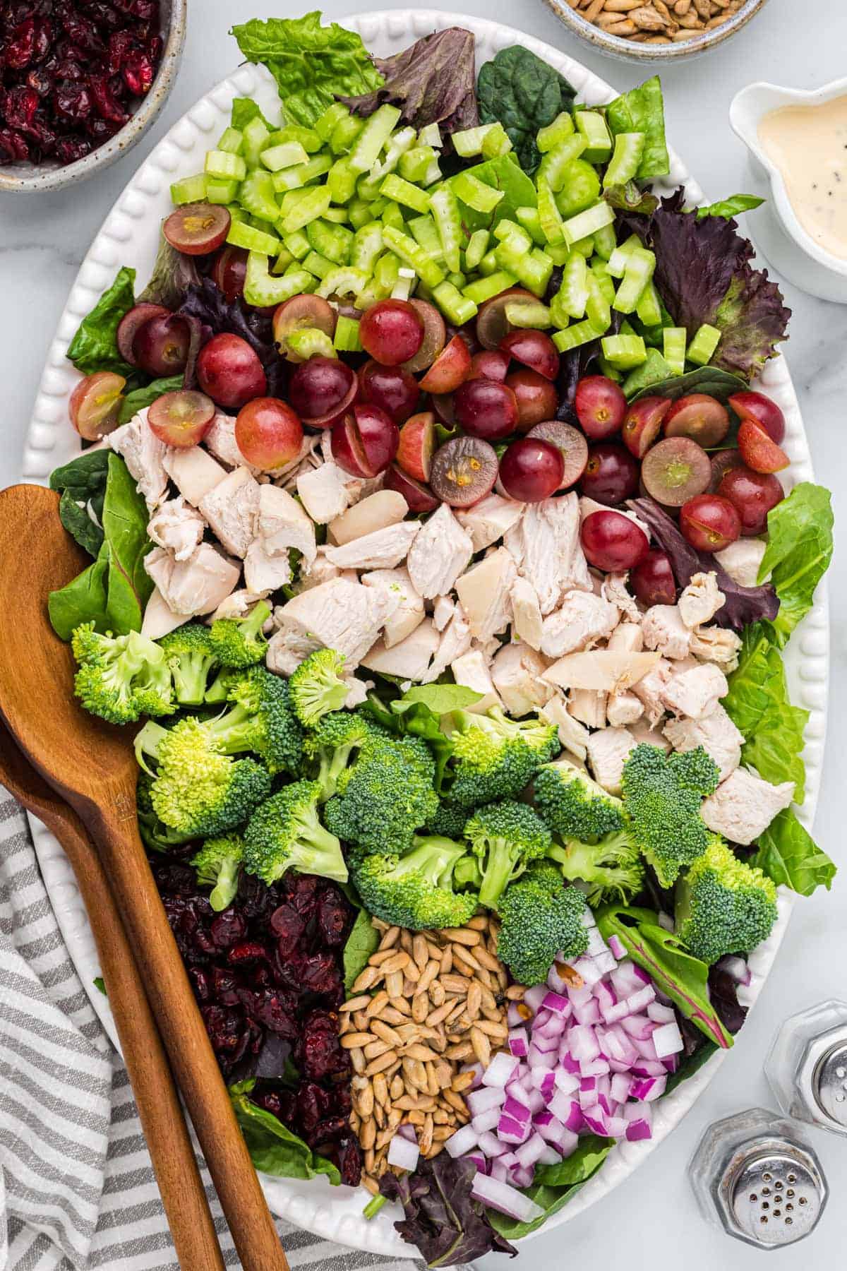 A fresh salad platter perfect for summer recipes, featuring mixed greens, celery, grapes, diced chicken, broccoli, red onion, sunflower seeds, and dried cranberries—served with wooden salad servers on the side.