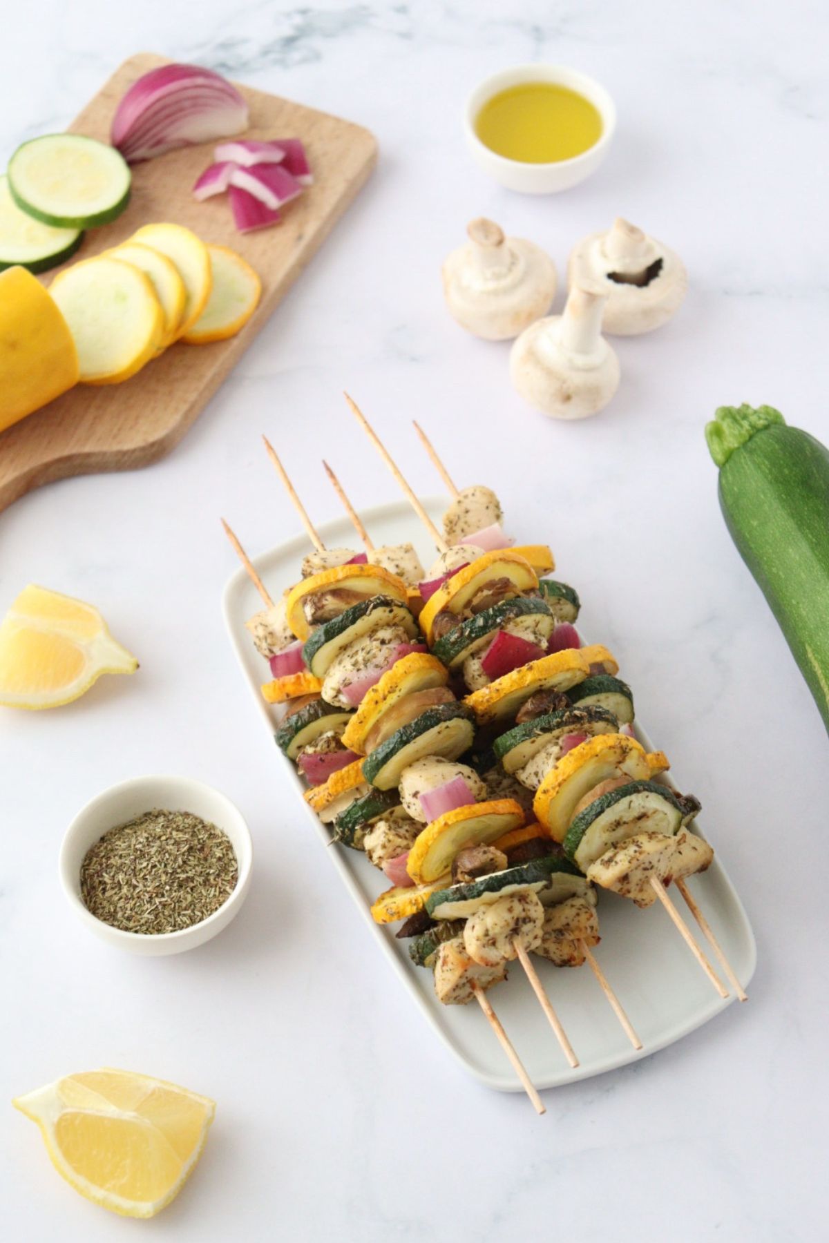 Four summer vegetable and grilled chicken skewers are arranged on a white platter, surrounded by fresh vegetables, lemon wedges, a bowl of herbs, and a small dish of olive oil on a white surface.
