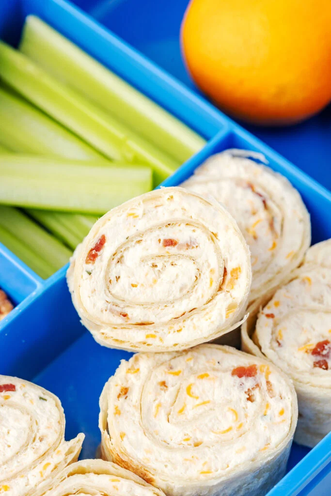 Pinwheel sandwiches with a creamy filling are packed in a blue lunchbox along with celery sticks and an orange—perfect for summer recipes or cold chicken recipes on the go.