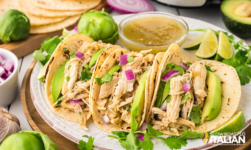 Three shredded chicken tacos with avocado slices, chopped red onion, and cilantro on corn tortillas are served on a white plate with salsa and lime wedges—perfect slow cooker fare for easy summer recipes. Fresh vegetables sit nearby.