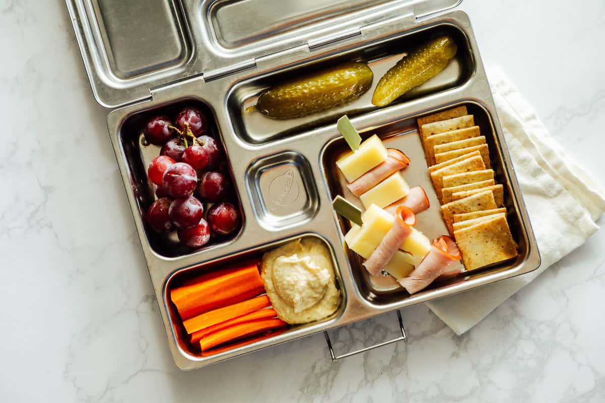A stainless steel lunchbox contains grapes, carrot sticks with hummus, a pickle, cheese and ham skewers, and a stack of crackers—perfect for kids lunches or as pool snacks on a sunny day.