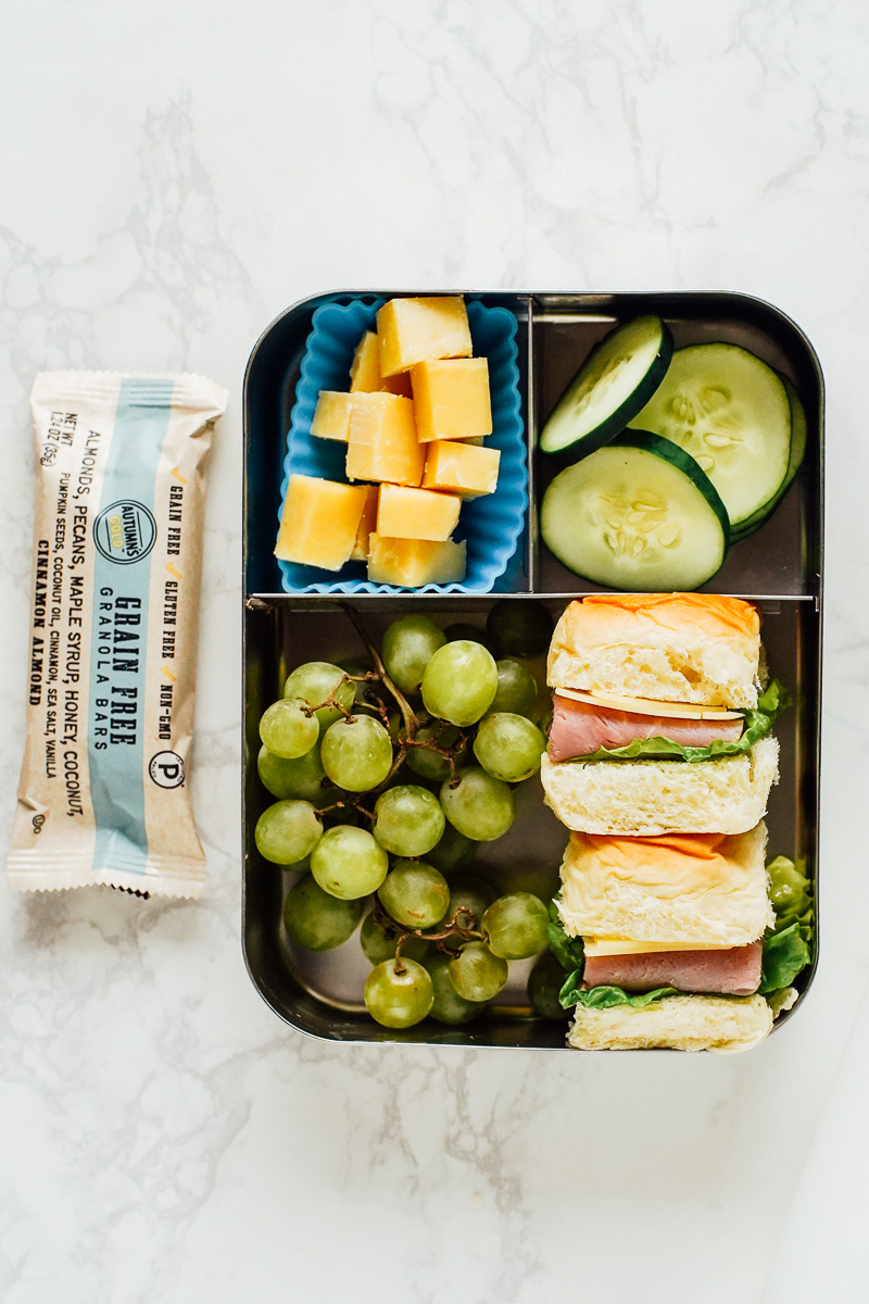 A lunchbox with cheese cubes, cucumber slices, grape clusters, mini sandwiches, and a packaged grain-free bar on a white surface—perfect cooler meals for picnic food or a relaxing boat day.