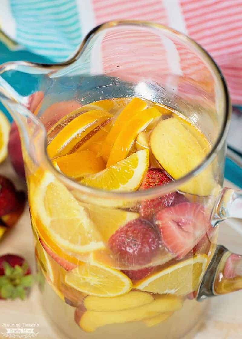 A glass pitcher filled with refreshing summer sangria, brimming with sliced oranges, lemons, strawberries, and ice.