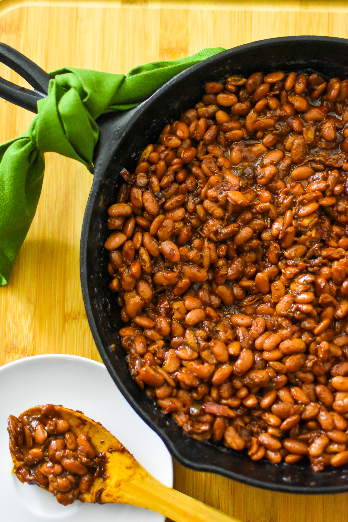 A cast iron skillet filled with cooked baked beans sits on a wooden surface, making these classic side dishes perfect for your next graduation party. A wooden spoon holds a portion of beans on a white plate beside it.