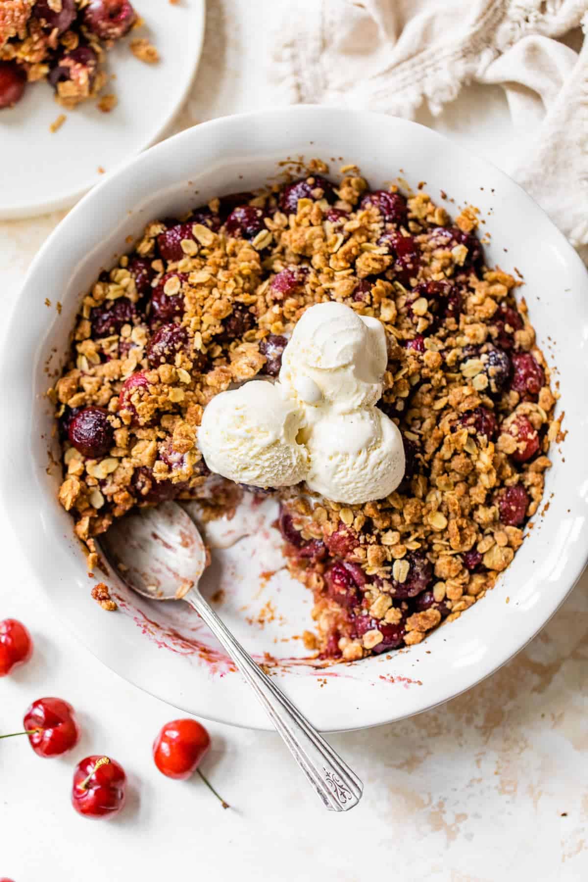 A cherry crisp, the perfect summer dessert, is topped with three scoops of vanilla ice cream in a white dish, with a serving spoon and fresh cherries nearby.