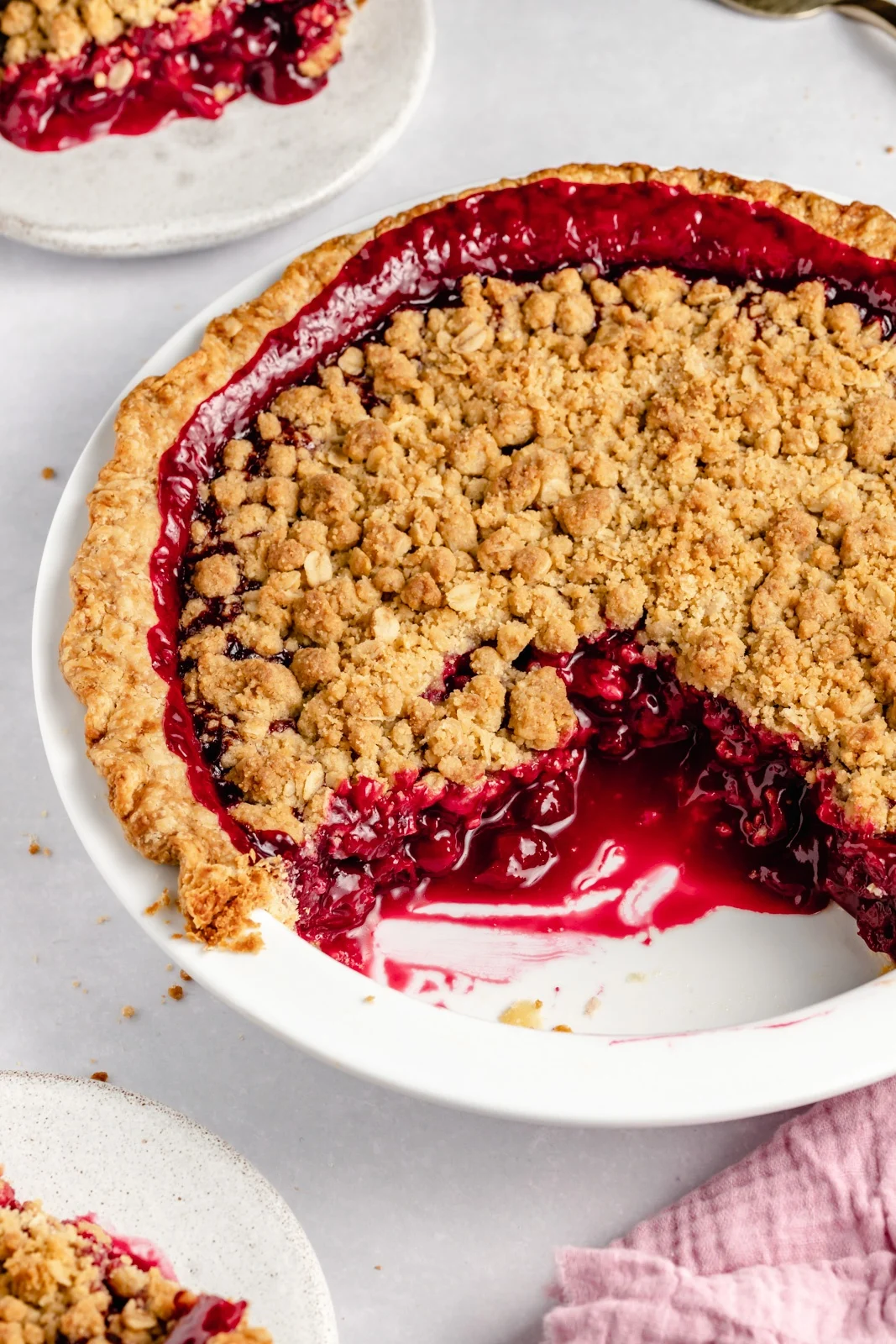 A fruit crumble pie with a golden crumb topping sits on a plate with a large slice missing, revealing a cherry-red berry filling—an inviting summer dessert.