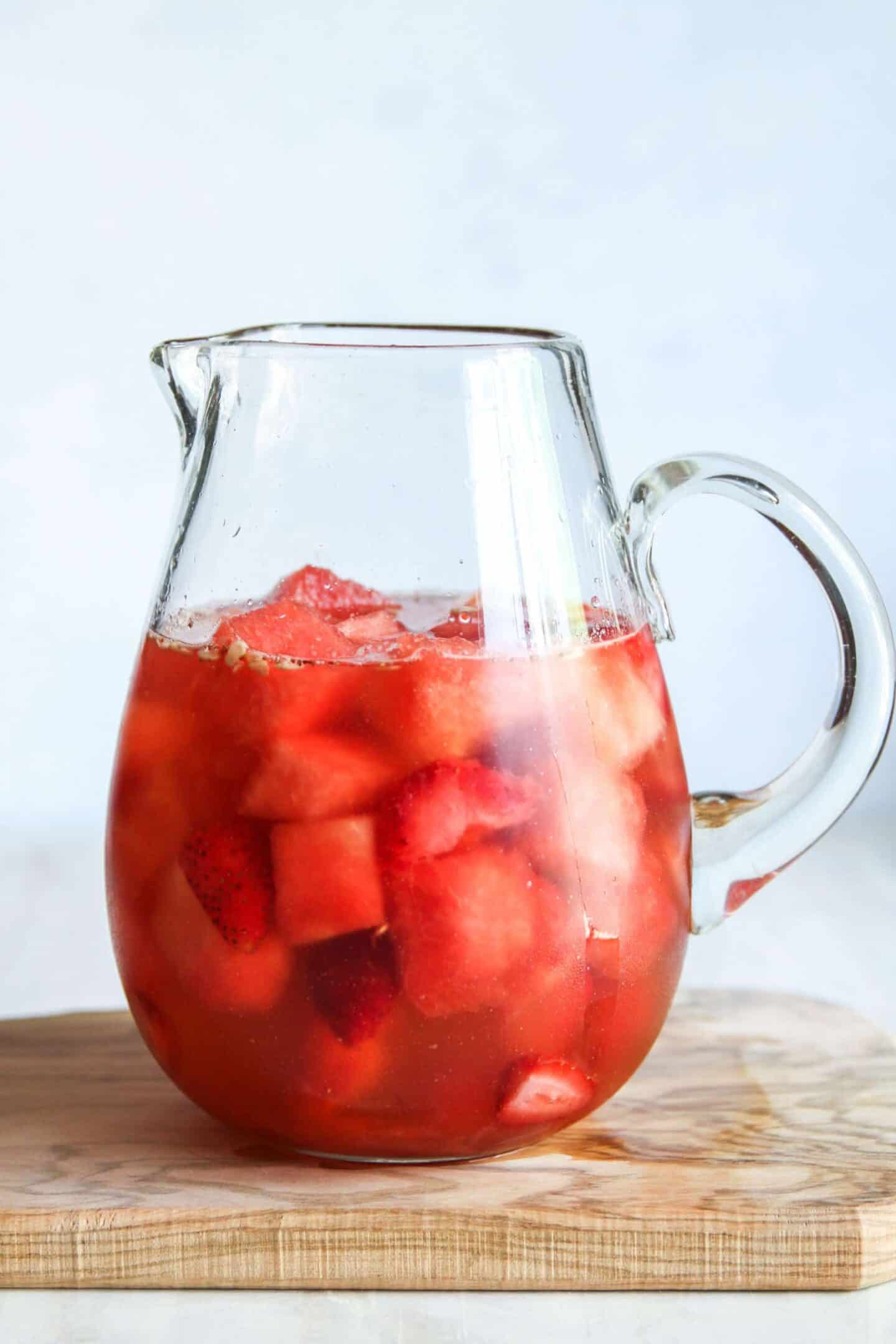 A clear glass pitcher filled with summer sangria, featuring a red fruit drink with chunks of strawberries and watermelon, placed on a wooden surface.