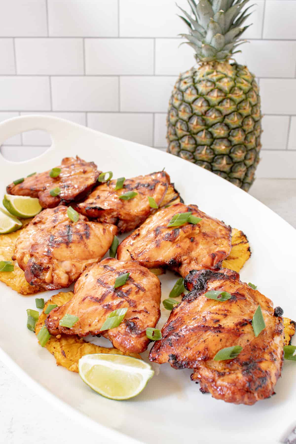 Grilled chicken thighs garnished with green onions and lime wedges, served on grilled pineapple slices with a whole pineapple in the background—a perfect dish for summer grilling.