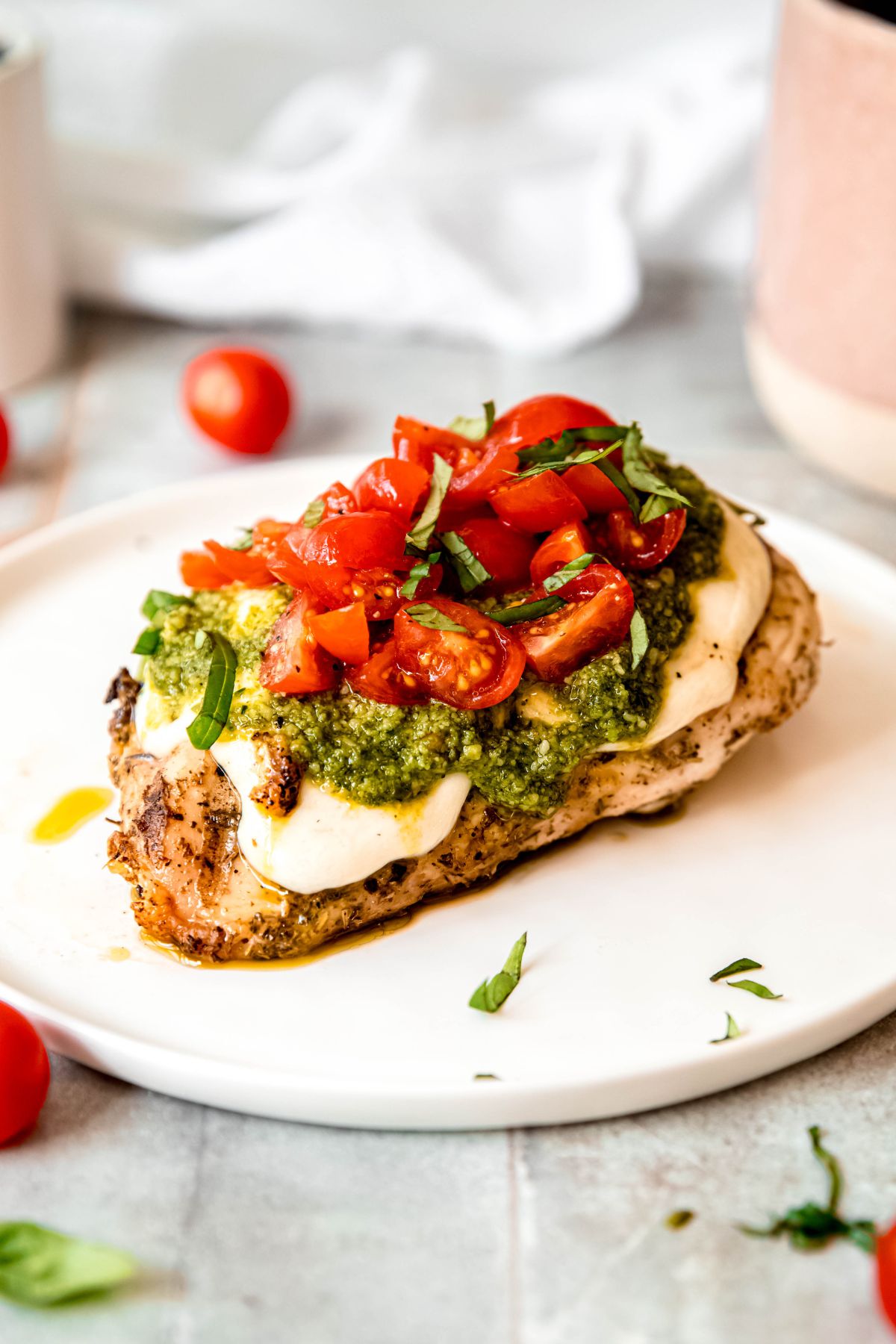 Grilled chicken breast topped with melted cheese, green pesto, chopped tomatoes, and fresh basil—an ideal choice for summer recipes—served on a white plate.