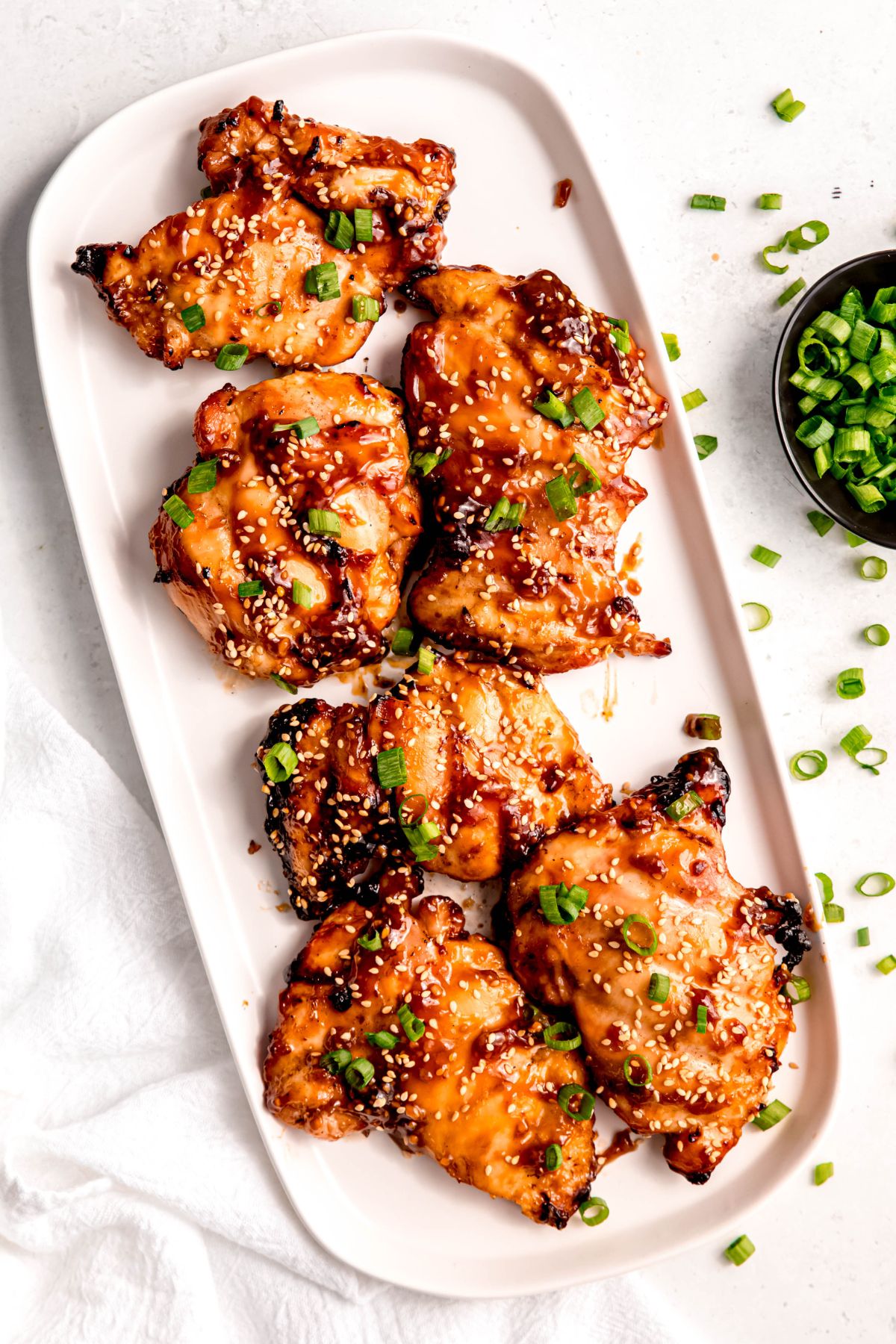 Summer grilled chicken thighs glazed with sauce, garnished with sesame seeds and chopped green onions, arranged on a white rectangular plate.