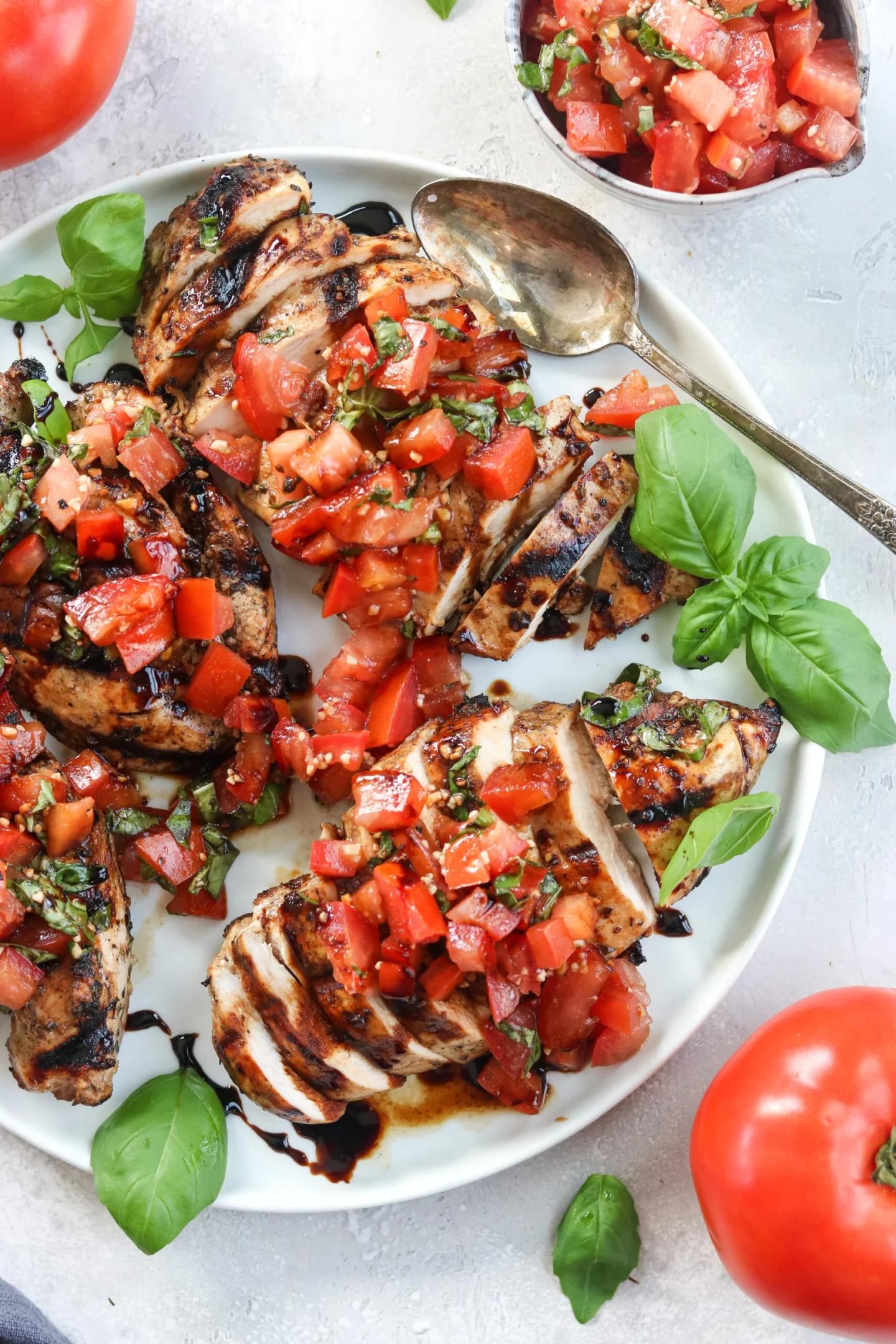 Summer grilled chicken breasts topped with chopped tomatoes, basil, and balsamic glaze, served on a white plate with fresh basil leaves and a spoon.