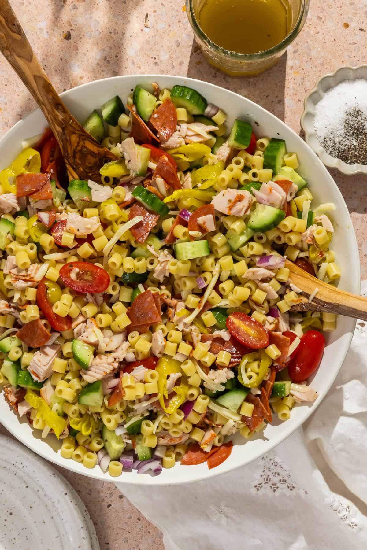 A bowl of pasta salad with chopped turkey, pepperoni, cucumber, cherry tomatoes, red onion, yellow peppers, and shredded cheese—perfect for beach lunch ideas or anyone wanting lunch without sandwiches. Dressing and pepper on the side with wooden serving utensils.