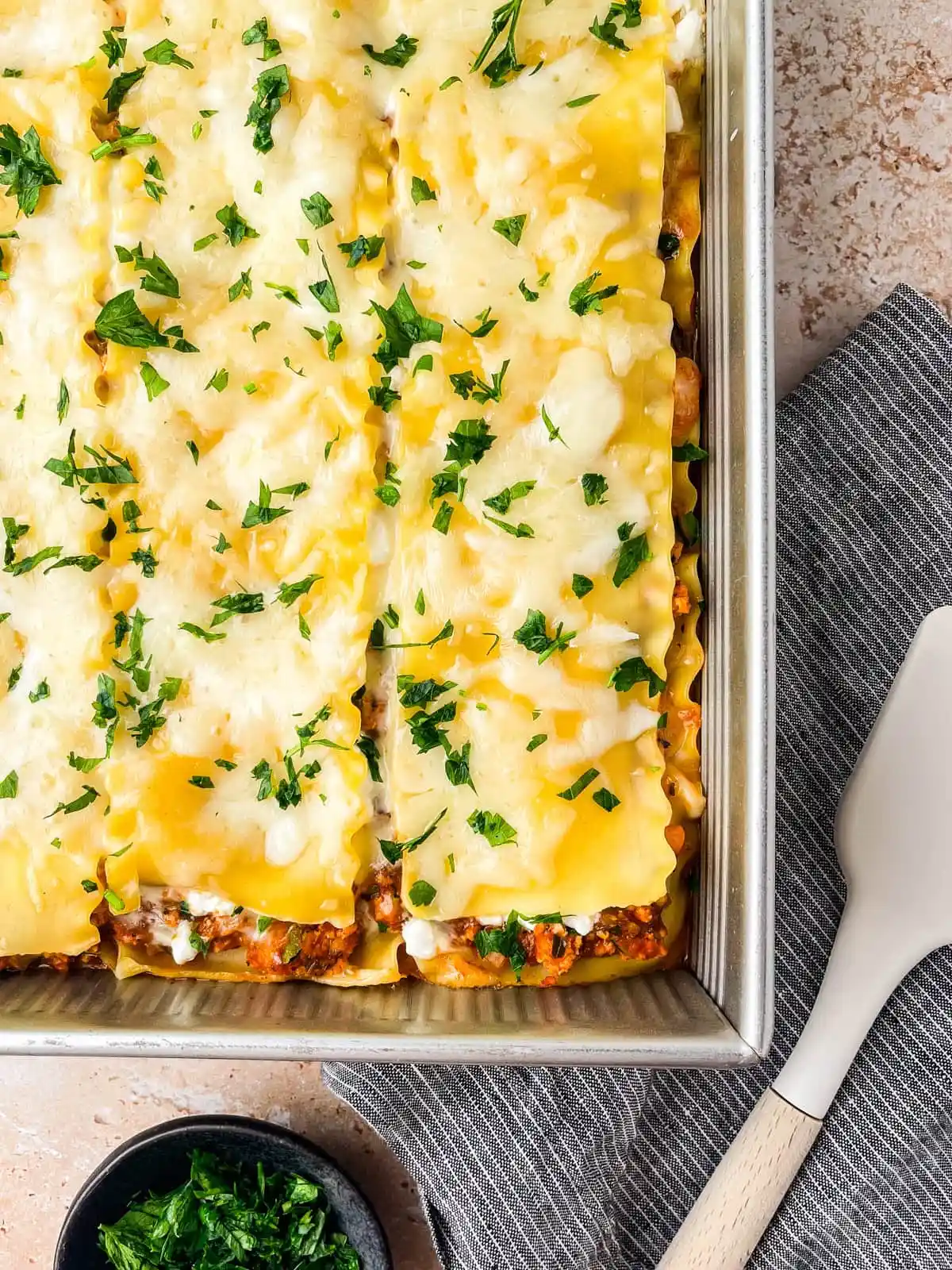 A baking dish with baked lasagna—one of the classic Italian Summer Recipes—topped with melted cheese and chopped parsley, next to a striped towel, white spatula, and a small bowl of parsley.