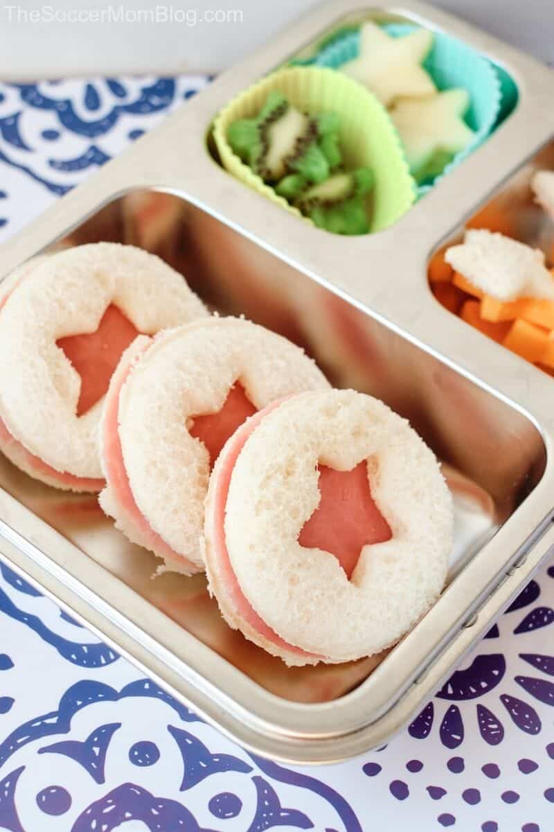 Three round sandwich halves with star-shaped cutouts are arranged in a metal lunch tray, with sliced fruit and veggies in separate compartments—a fun kids lunch perfect for pool snacks or creative pool lunch ideas.