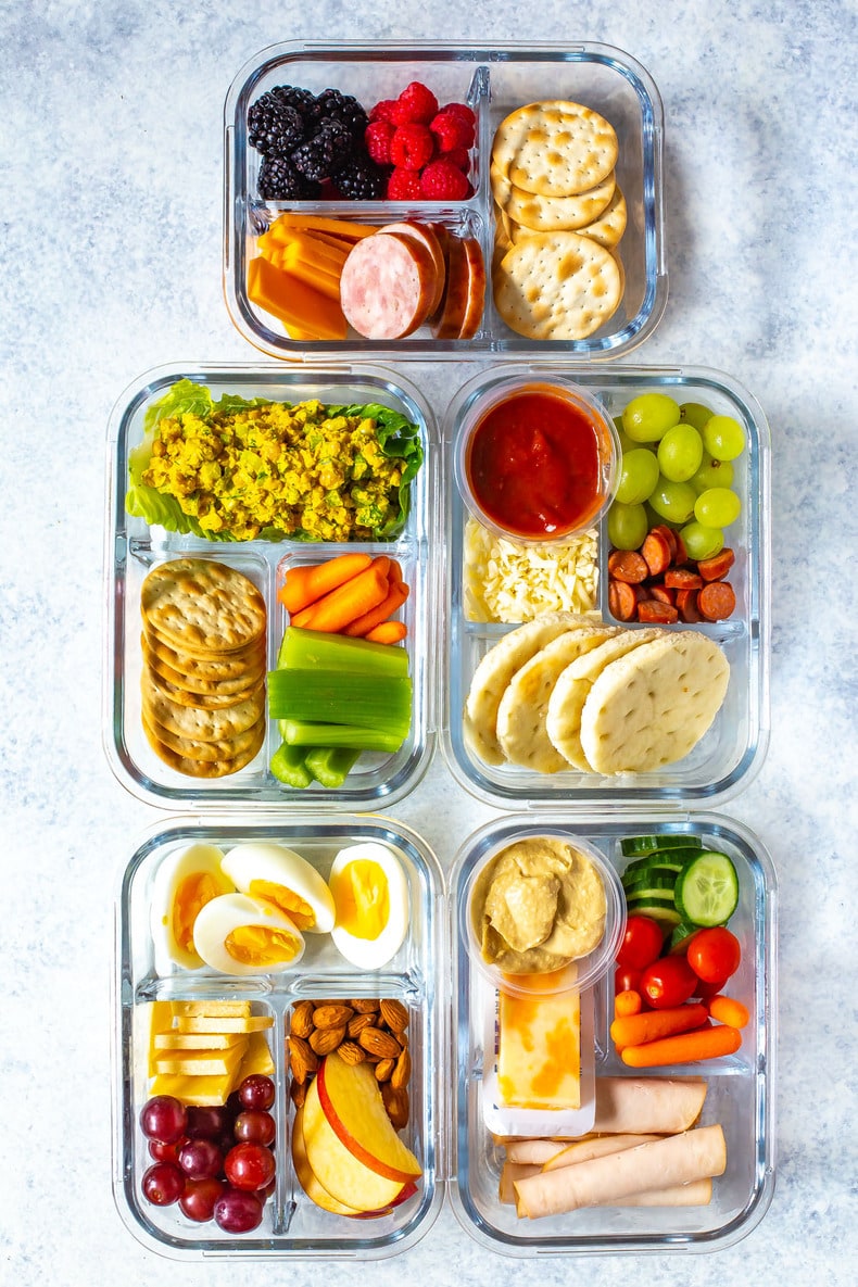 Five meal prep containers filled with assorted snacks—perfect boat food for a boat day—including crackers, fruits, vegetables, boiled eggs, hummus, cheese, nuts, lunch meats, and sauces. Great cooler meals for your adventure!.