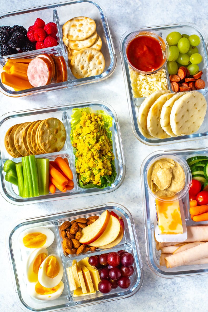 Five meal prep containers filled with a variety of snacks—crackers, fruit, veggies, cheese, nuts, hummus, and boiled eggs—are neatly arranged in partitions; perfect for cold beach lunch or easy beach lunch ideas.