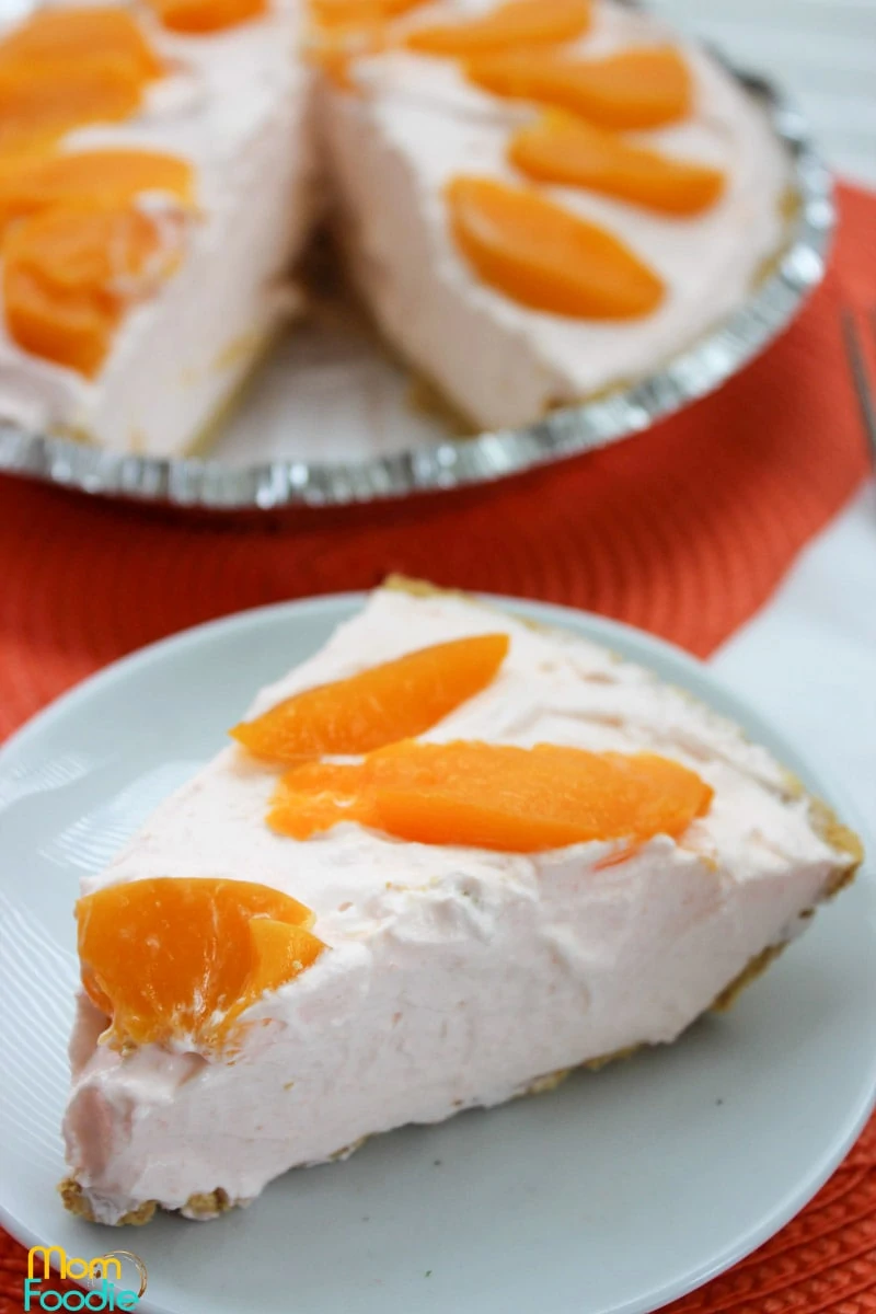 A slice of creamy pie topped with orange fruit slices sits on a plate in front of the remaining peach dessert.