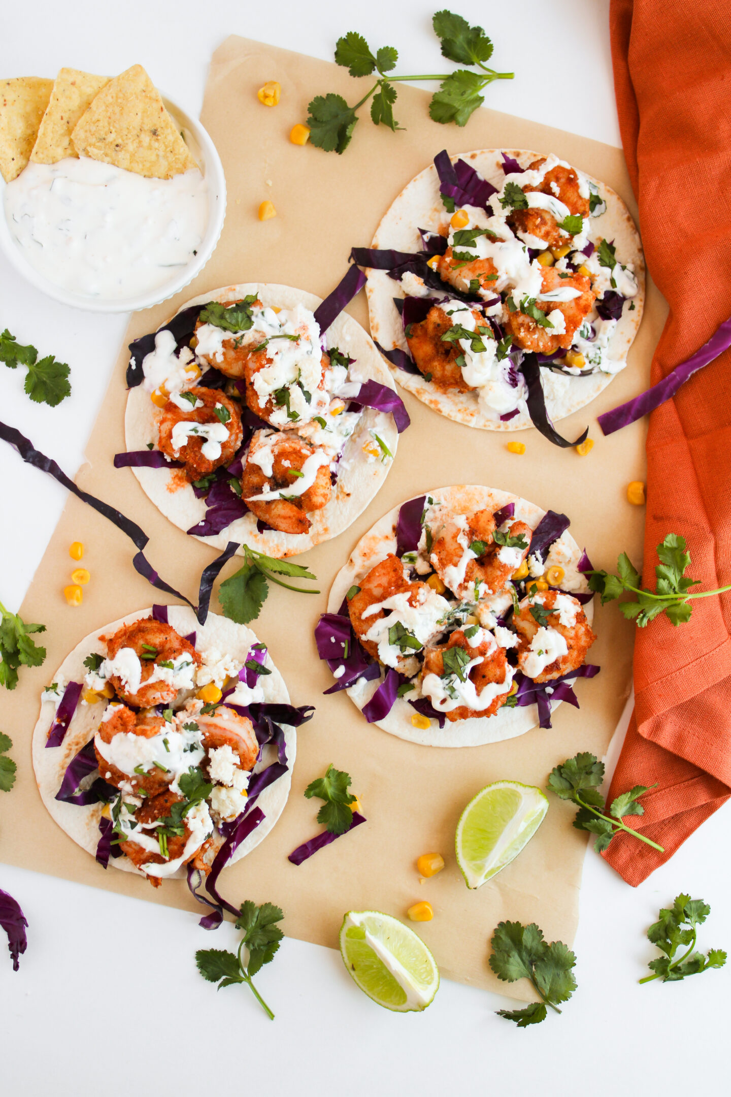 Four tacos topped with buffalo cauliflower, shredded purple cabbage, white sauce, cilantro, and cheese on parchment paper—perfect for a summer dinner—surrounded by lime wedges, cilantro, tortilla chips, and an orange napkin.