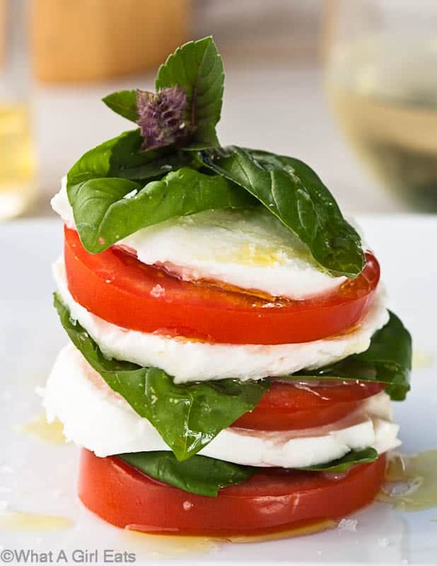 Stacked slices of tomato, mozzarella, and basil leaves, drizzled with olive oil and garnished with a sprig of basil on a white plate—an irresistible choice for Italian Summer Recipes.
