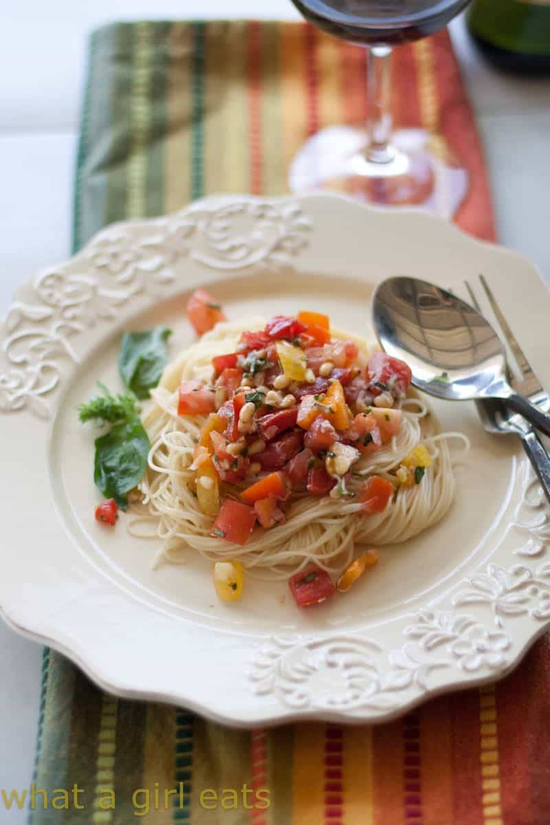 A plate of angel hair pasta topped with a fresh tomato and vegetable sauce, garnished with herbs, makes a perfect choice for Italian Summer Recipes, served beside a spoon and fork on a patterned placemat.