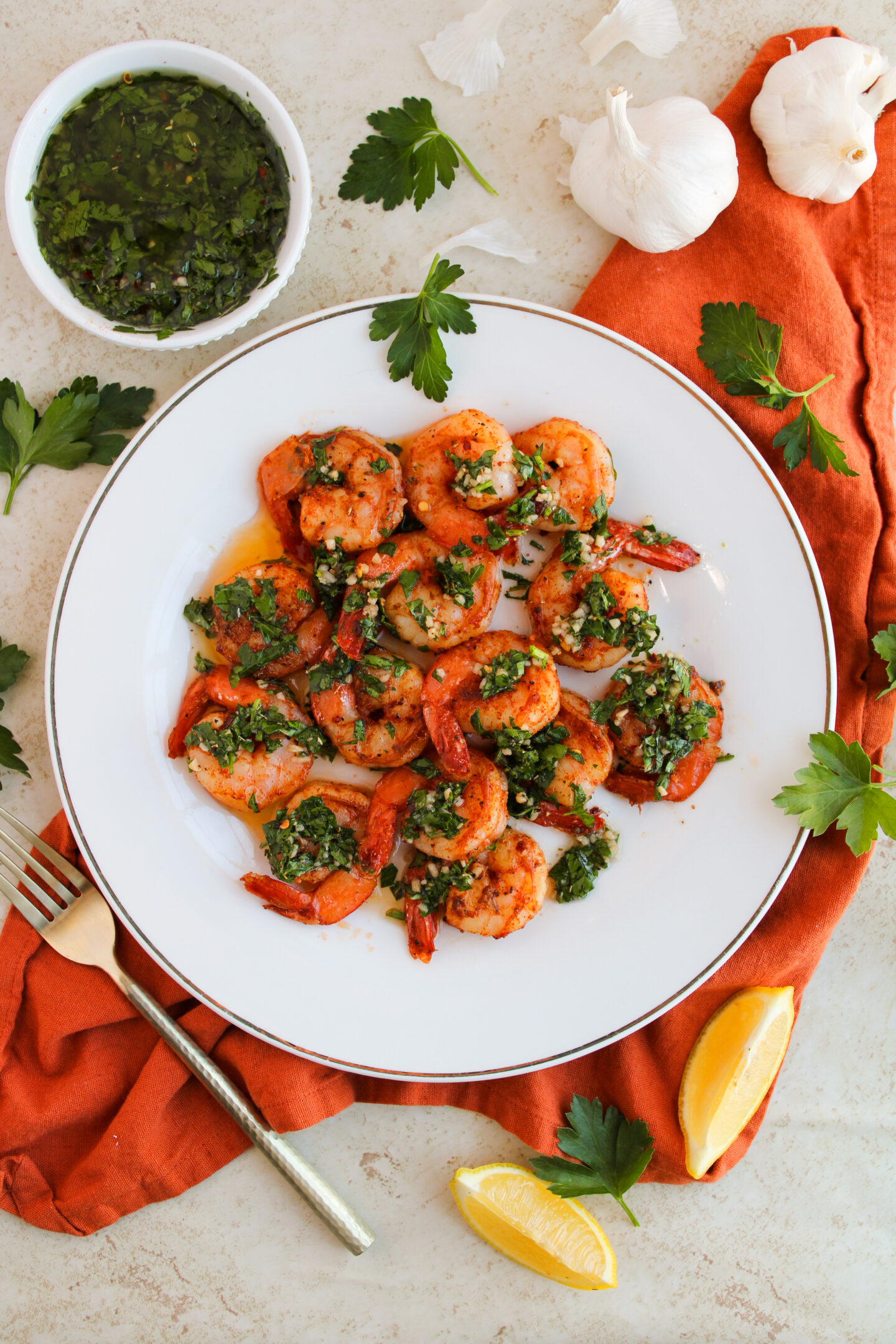 A plate of cooked shrimp topped with green herb sauce and garnished with parsley makes a perfect summer dinner. Lemon wedges, a fork, and a bowl of sauce sit nearby on an orange napkin.