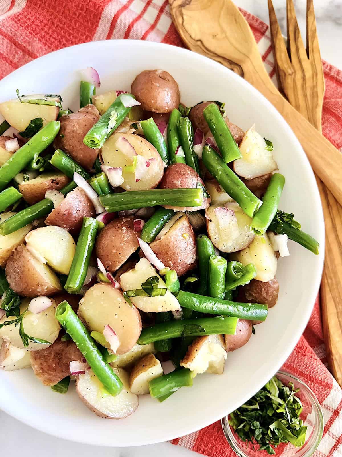 A white bowl filled with a potato salad of red potatoes, green beans, red onions, and herbs sits on a red and white cloth with wooden serving utensils—perfect for Italian Summer Recipes.