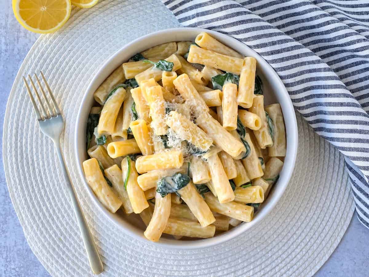 A bowl of rigatoni pasta with creamy sauce, spinach, and grated cheese on top, perfect for Italian Summer Recipes, is placed on a white placemat with a fork, striped napkin, and lemon slices nearby.