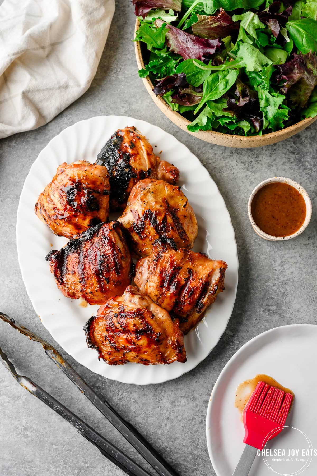 Summer meets flavor with grilled chicken thighs on a white plate, served alongside a bowl of mixed greens and dressing. Metal tongs and a red basting brush add the final touch to this fresh, seasonal meal.