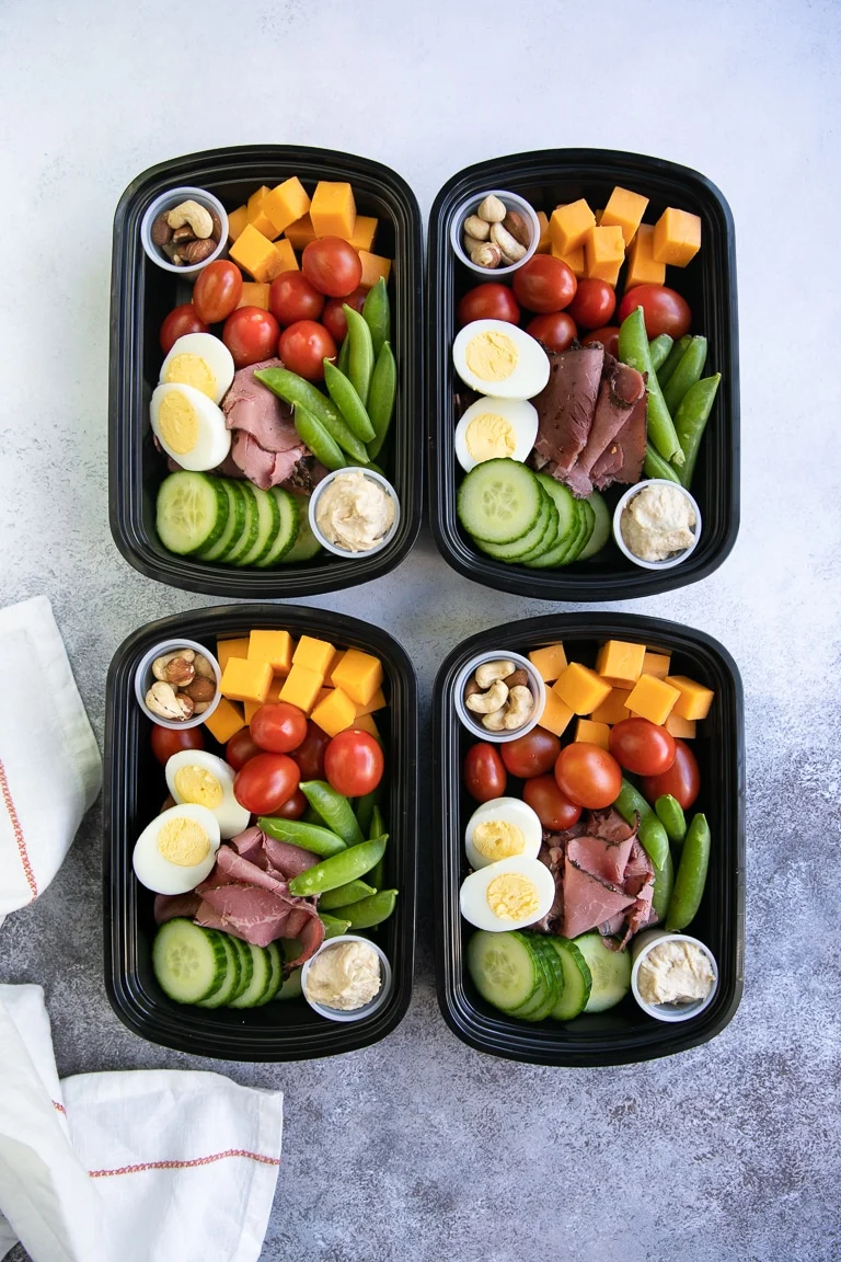 Four black meal prep containers filled with sliced boiled eggs, roast beef, cucumber, sugar snap peas, cherry tomatoes, cheddar cheese cubes, nuts, and a small container of dip—perfect cold lunch ideas for a tasty beach lunch.