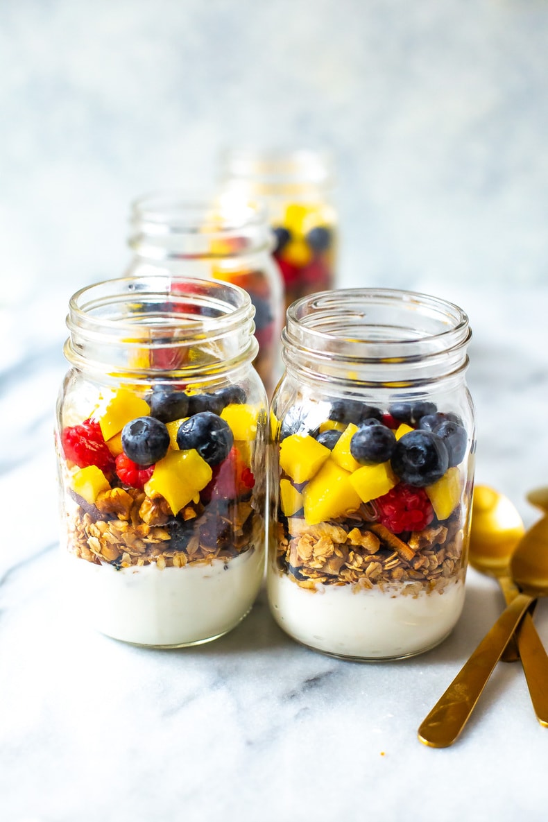 Four glass jars filled with layers of yogurt, granola, mango, blueberries, and raspberries are arranged on a marble surface with gold spoons—perfect for cold lunch ideas or a refreshing cold beach lunch.