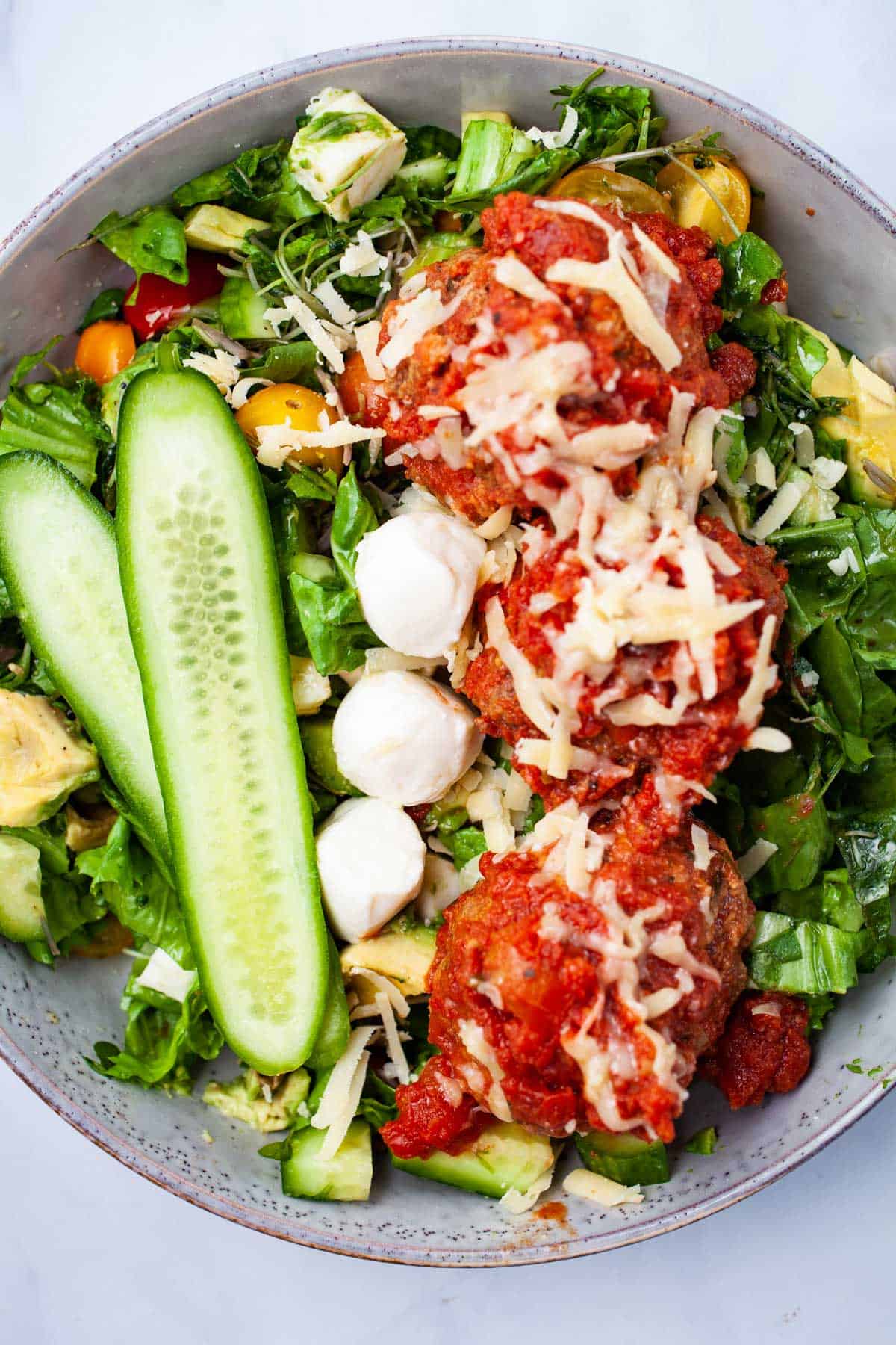 A bowl of salad topped with three meatballs in tomato sauce, shredded cheese, sliced cucumber, mini mozzarella balls, and mixed vegetables—perfect for those seeking delicious Italian Summer Recipes.