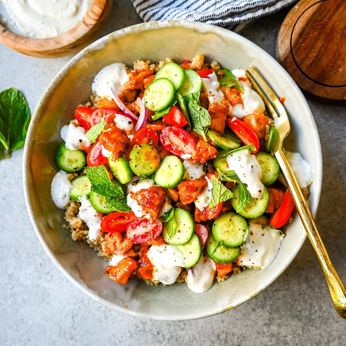 A refreshing summer salad with cucumbers, tomatoes, red onions, fresh mint, quinoa for high protein, and creamy white dressing, served with a gold fork on a gray surface.