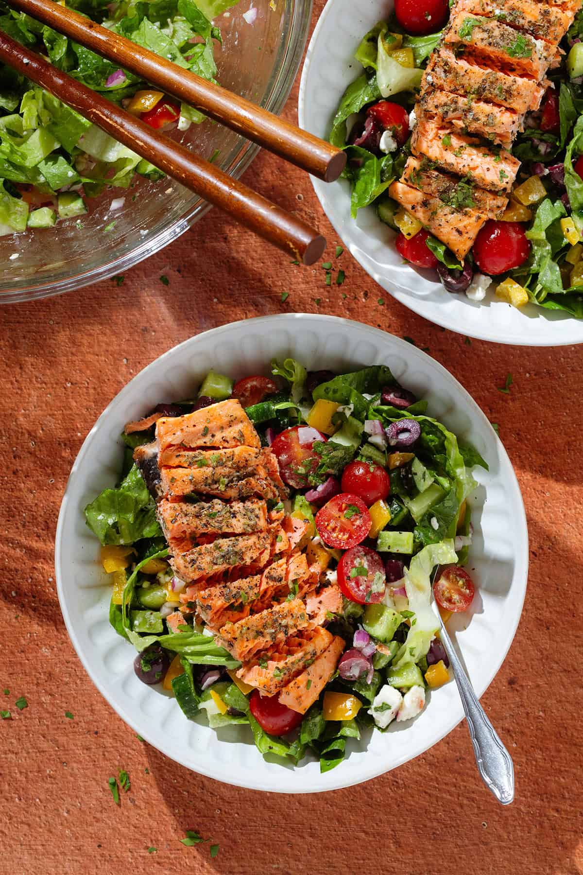 Two bowls of high protein salad topped with grilled salmon, cherry tomatoes, olives, and feta cheese make for a perfect summer meal, with a glass bowl of salad and chopsticks in the background.