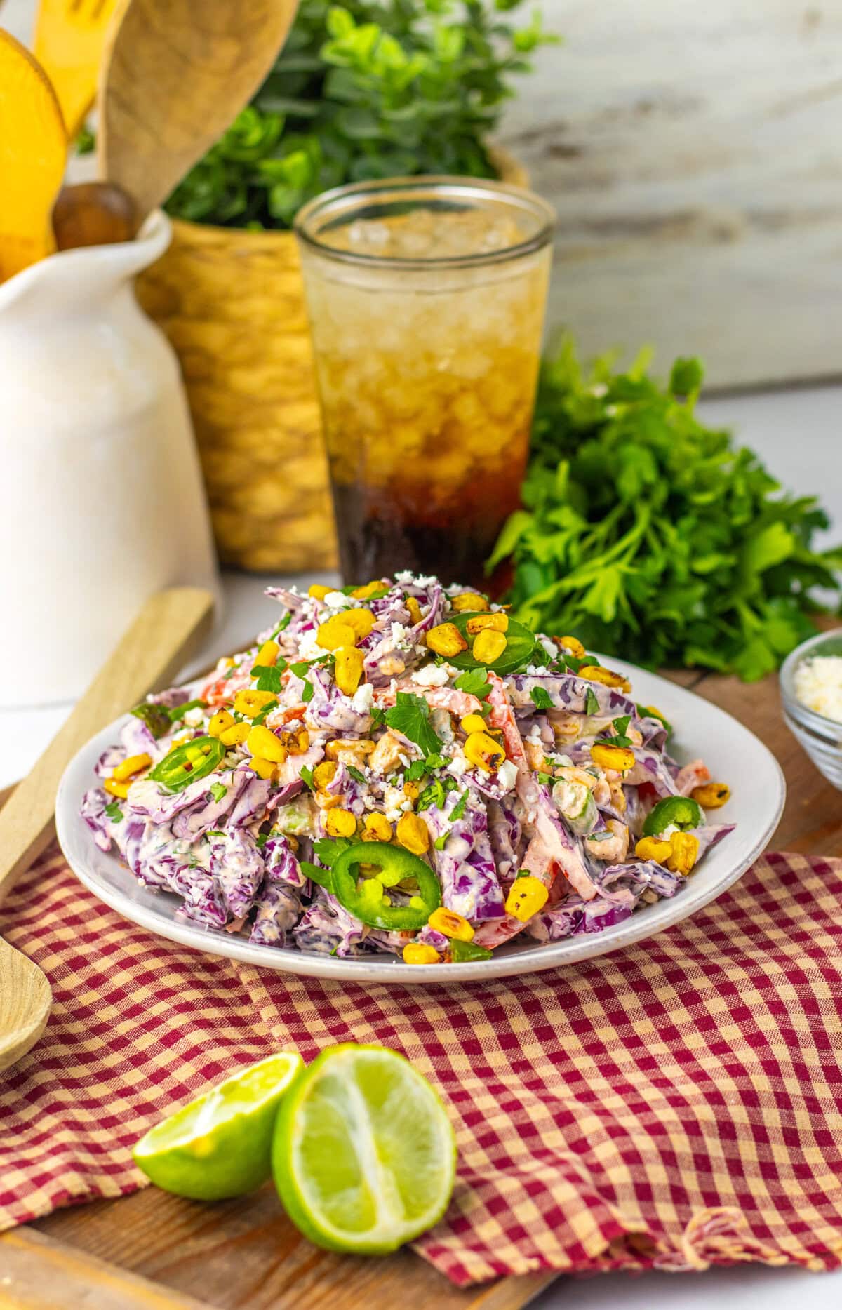A plate of colorful coleslaw, perfect for side dishes at a graduation party, is topped with sliced jalapeños, crumbled cheese, and corn. A cut lime, wooden spoon, and a glass of iced drink rest in the background.