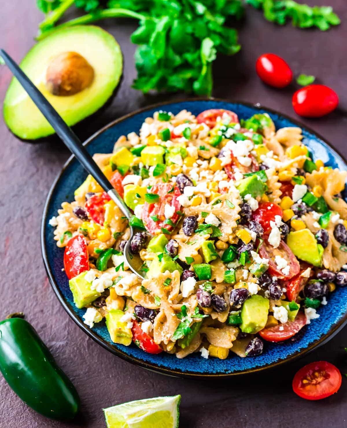 A bowl of pasta salad with black beans, avocado, cherry tomatoes, corn, green onions, crumbled cheese, and a fork—perfect for Easy Summer Recipes. Surrounding ingredients include jalapeño, lime, avocado, and cilantro.