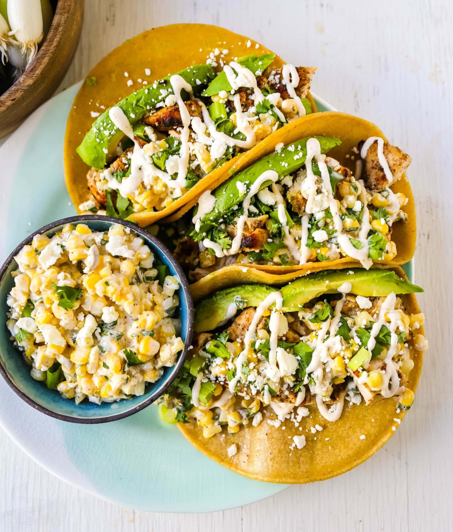 Three high protein tacos with avocado, chicken, cheese, and sauce are served on a plate next to a bowl of corn salad topped with crumbled cheese—perfect for a summer meal.