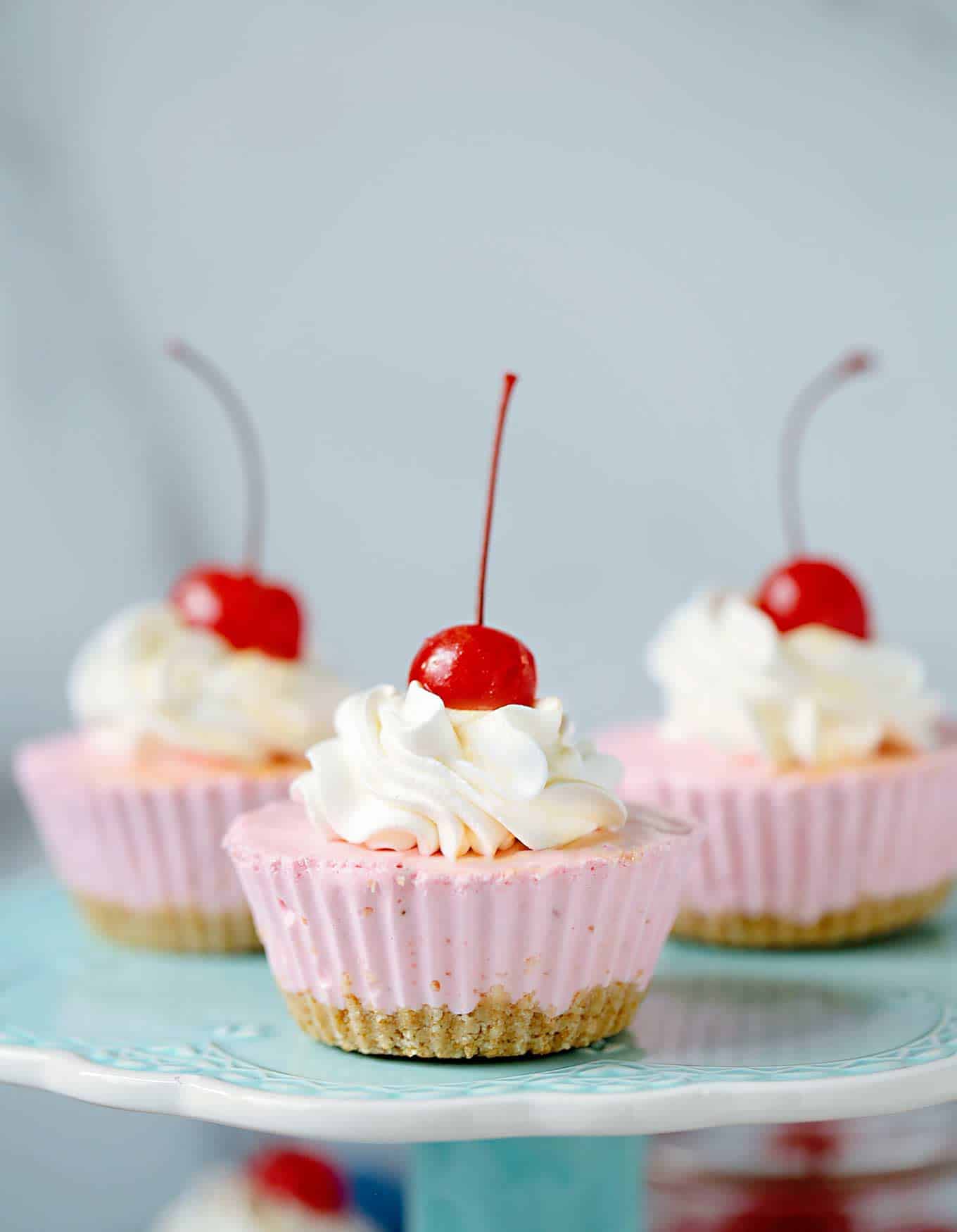 Three pink mini cheesecake cupcakes with whipped cream and cherries on top, displayed on a light blue cake stand—a perfect cherry-filled summer dessert.