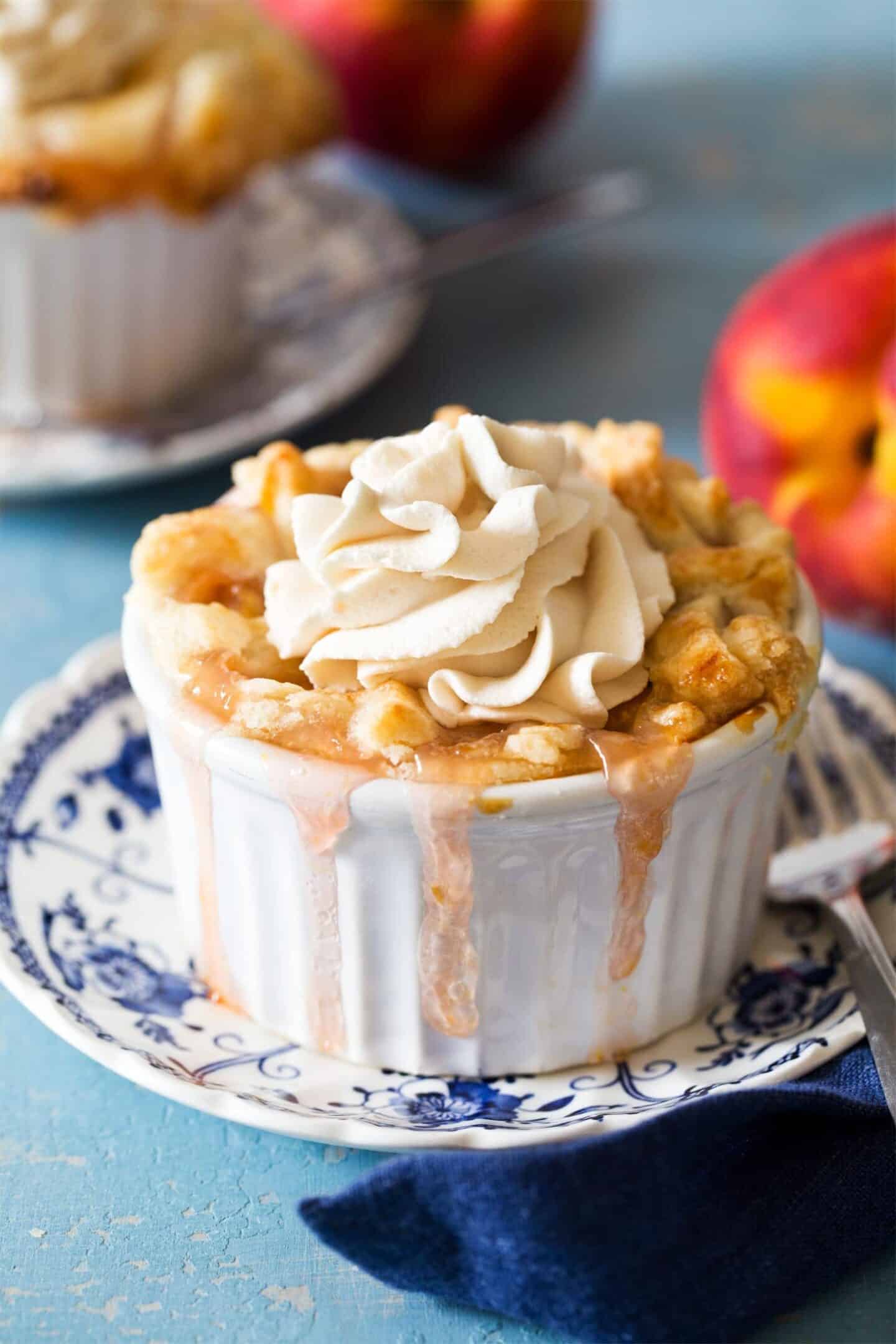 A small peach dessert cobbler in a white ramekin topped with whipped cream, with filling dripping over the sides, sits on a decorative plate. Peaches are visible in the background.
