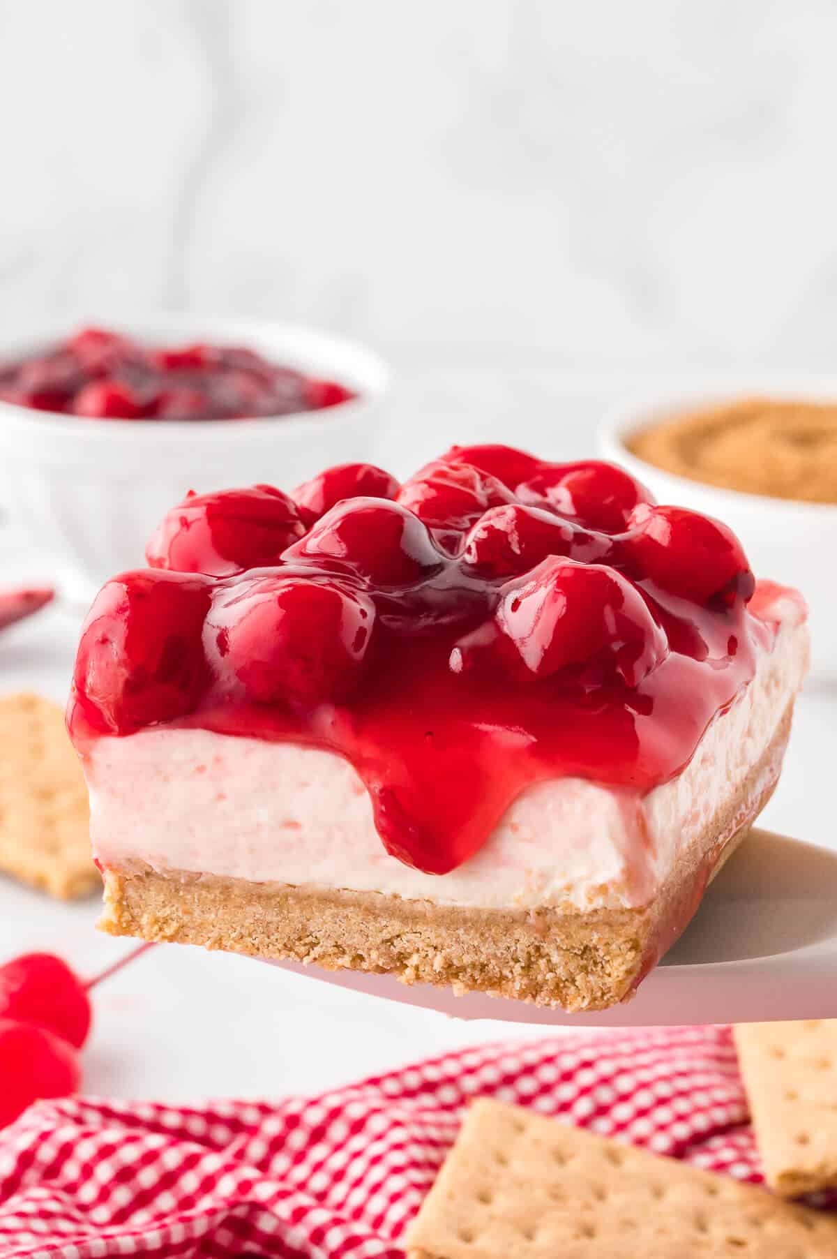 A slice of cherry-topped cheesecake with a graham cracker crust is being lifted from a tray, making it the perfect summer dessert, with graham crackers and bowls of ingredients in the background.