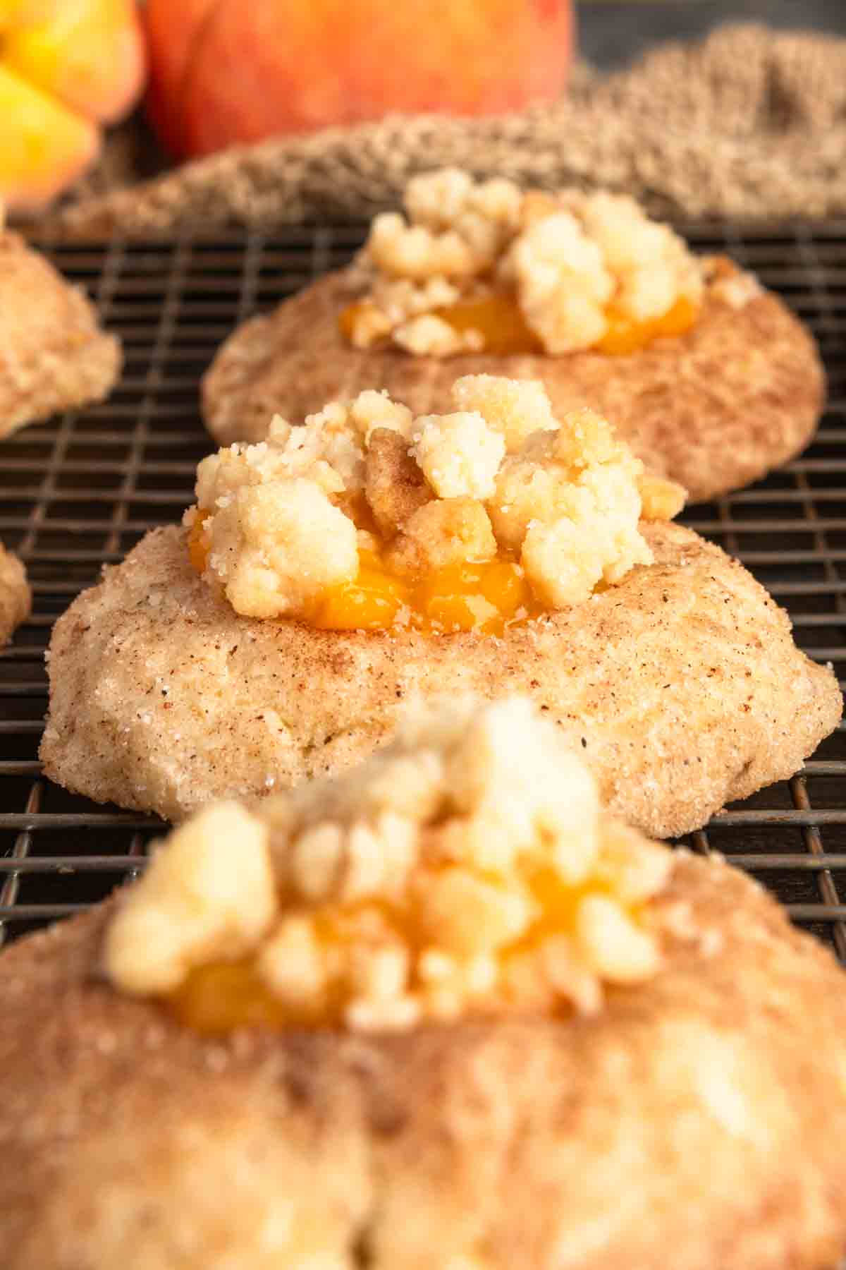 Close-up of cookies topped with crumbly streusel and peach dessert filling, cooling on a wire rack with apples and a cloth in the background.