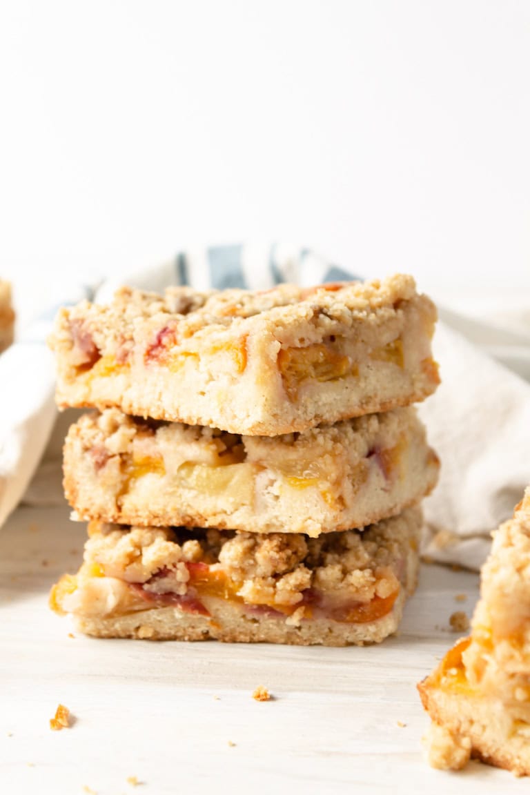 A stack of three peach crumb bars, a delicious peach dessert, sits on a light surface with crumbs scattered around and a cloth in the background.