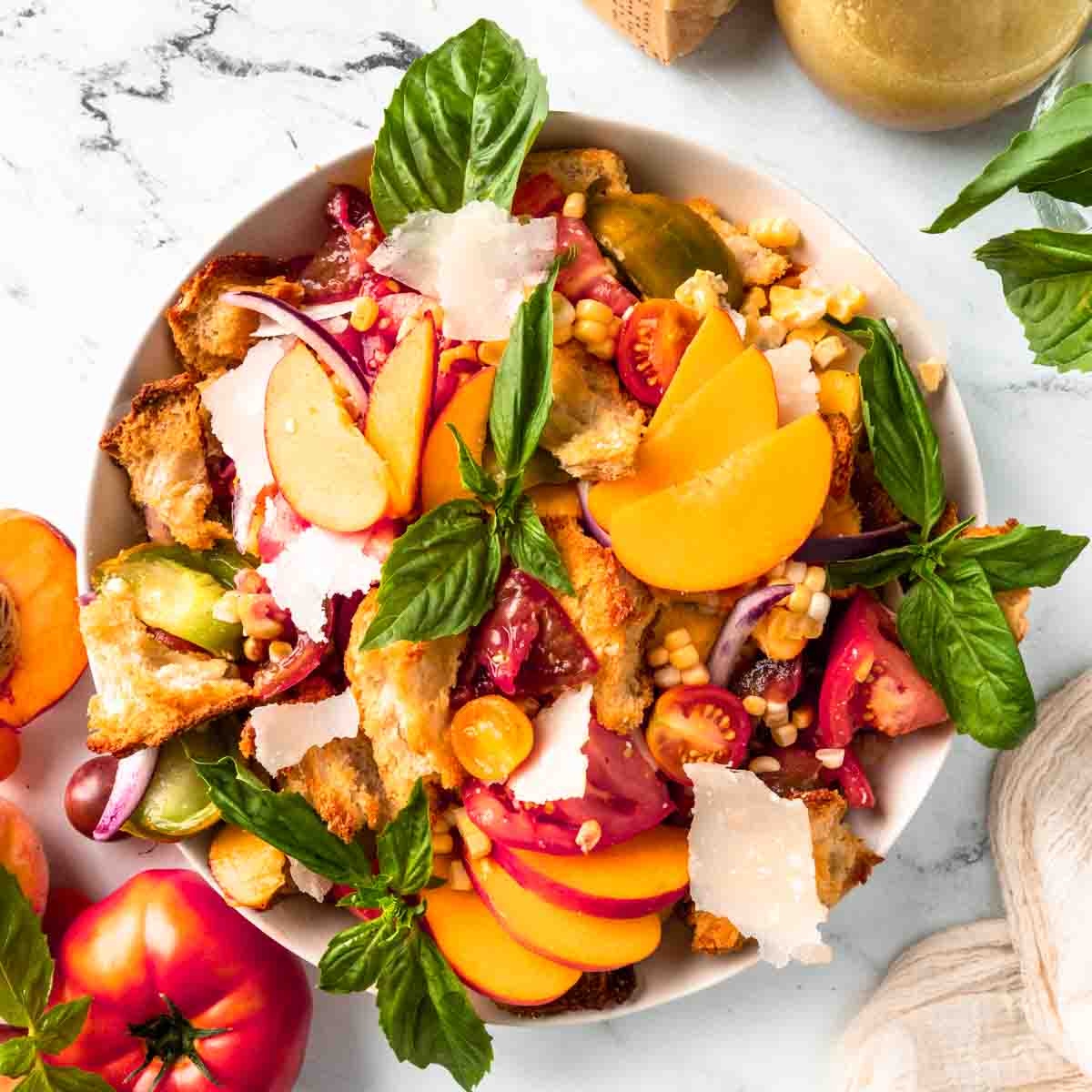 A bowl of panzanella salad featuring tomatoes, peaches, red onions, corn, croutons, basil leaves, and shaved cheese on a marble surface—perfect for Italian Summer Recipes.