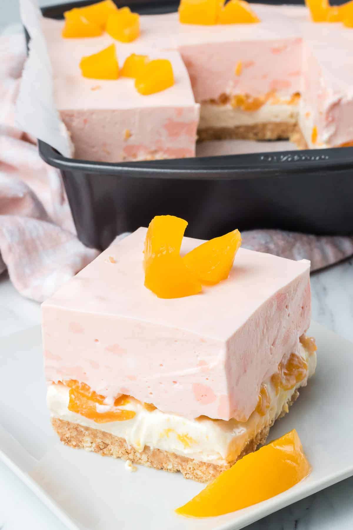 A square slice of layered peach dessert with a pink creamy top, peach pieces, and a crumbly base sits on a plate with extra peach pieces. The rest of the peach dessert remains in a baking dish nearby.