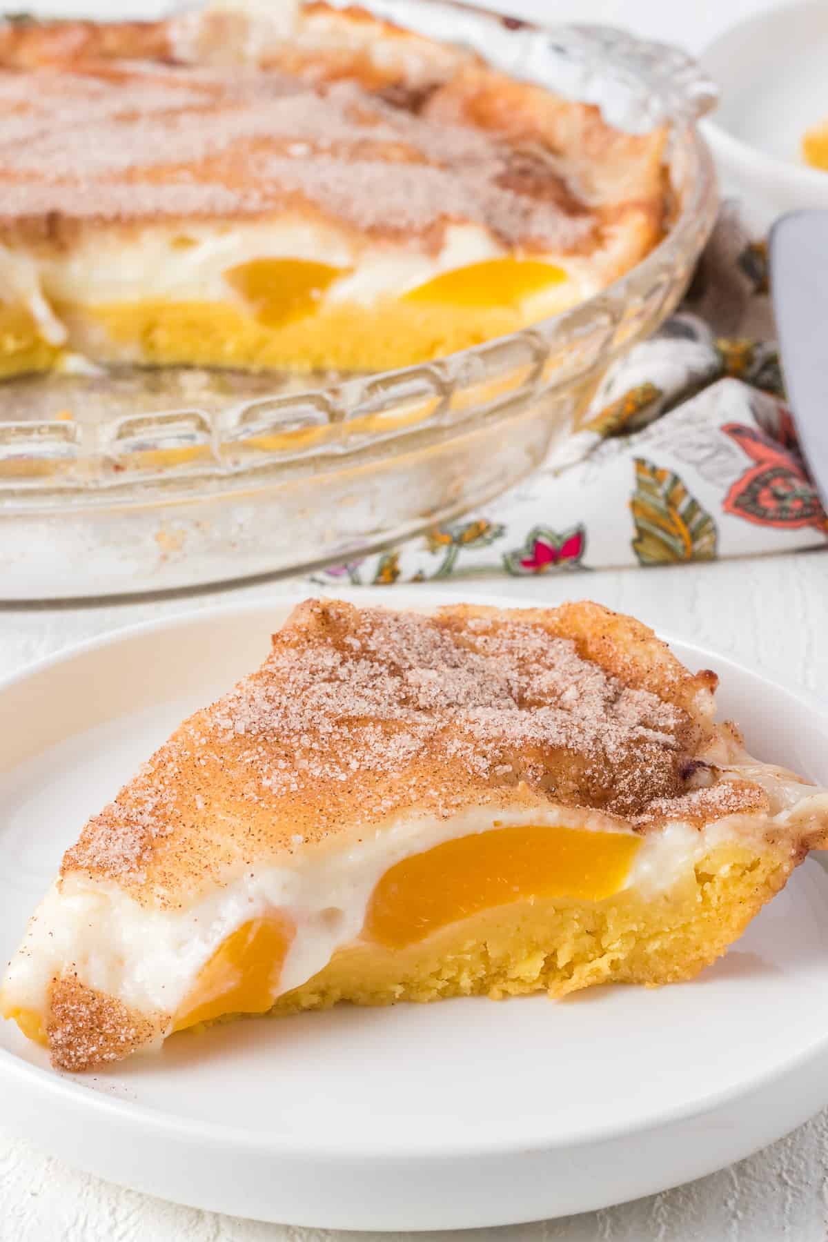 A slice of peach custard pie, a classic peach dessert, topped with cinnamon sugar on a white plate, with the rest of the pie in a glass dish in the background.