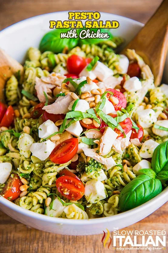 A bowl of pesto pasta salad with chicken, cherry tomatoes, mozzarella balls, pine nuts, and fresh basil—perfect as picnic food or for those searching for tasty cold beach lunch ideas. Wooden serving utensils complete the summery vibe.