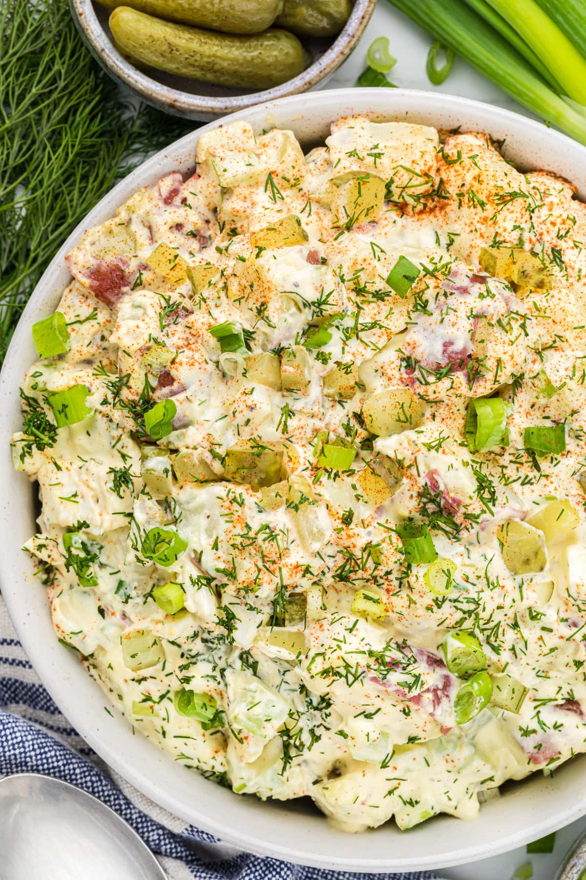 A bowl of creamy potato salad garnished with chopped dill, green onions, and sliced pickles—an irresistible side dish perfect for any graduation party, served alongside a bowl of pickles and fresh herbs in the background.