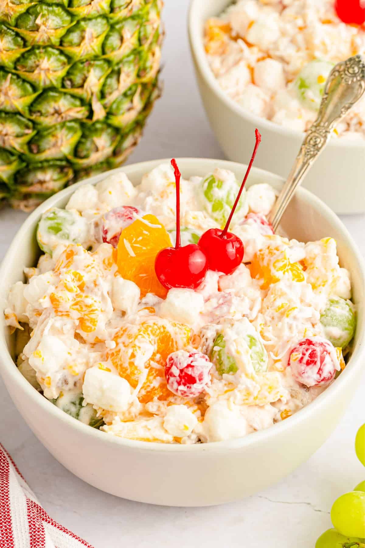 A bowl of fruit salad with marshmallows, grapes, mandarin oranges, pineapple, and cherries—perfect for side dishes at a graduation party—topped with two maraschino cherries and set beside a fresh pineapple.