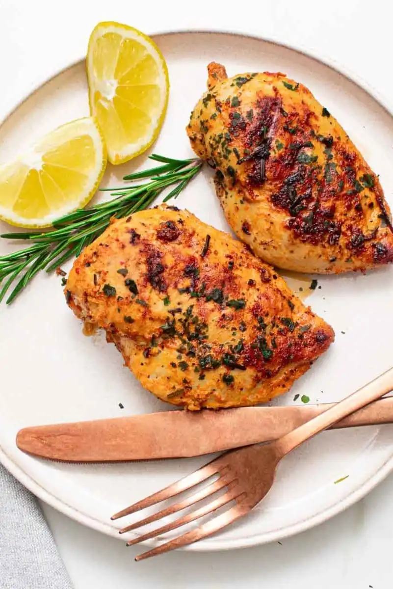 Two grilled chicken breasts garnished with herbs sit on a plate with two lemon wedges, a sprig of rosemary, and a fork and knife—perfect for a light summer meal.