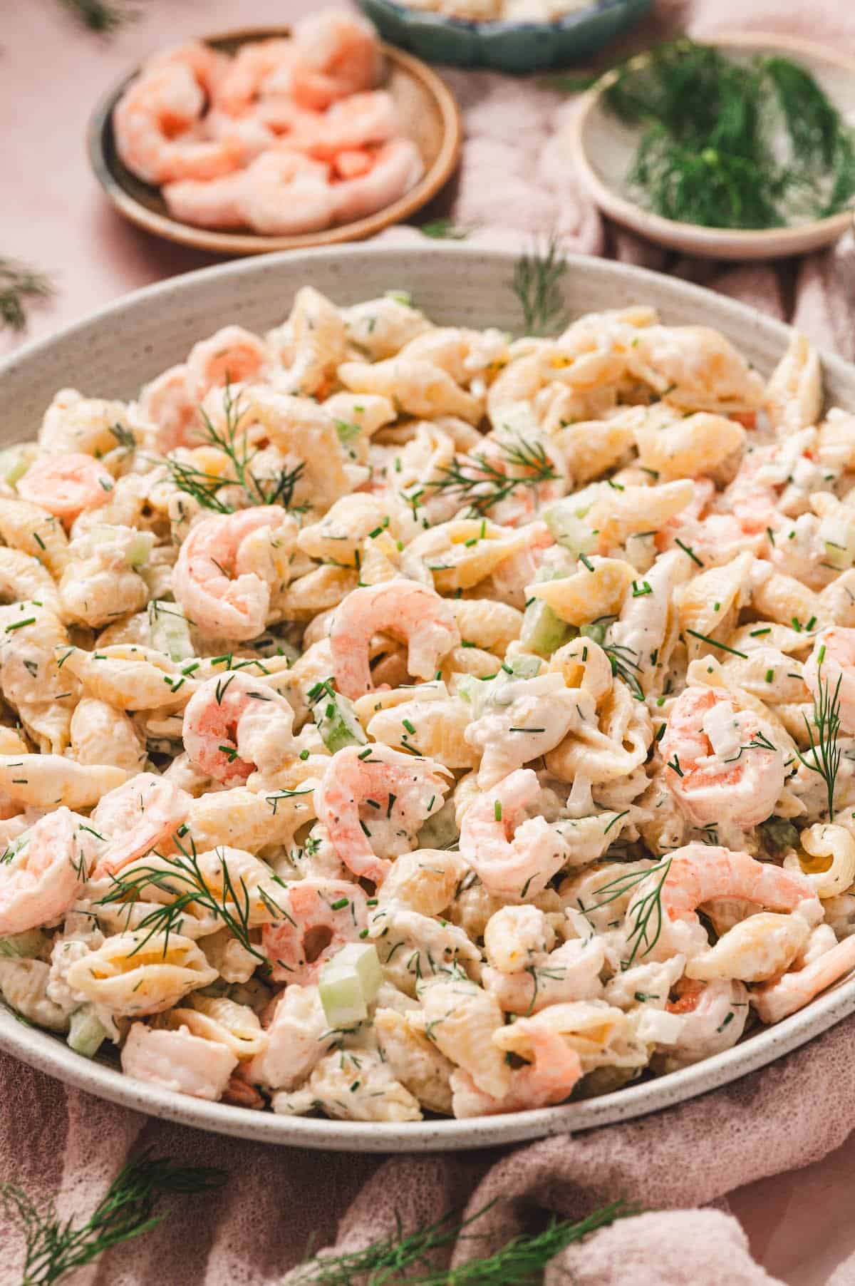 A large bowl of creamy pasta salad with shrimp, diced celery, and fresh dill, perfect as one of the side dishes for a graduation party, surrounded by small bowls of shrimp and dill on a table.